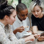 Drei junge Menschen sitzen gemeinsam an einem Tisch, schauen auf ein Smartphone und unterhalten sich. Eine von ihnen hält eine Tasse Kaffee.
