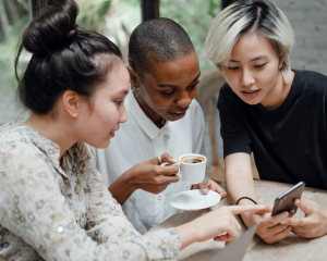 Drei junge Menschen sitzen gemeinsam an einem Tisch, schauen auf ein Smartphone und unterhalten sich. Eine von ihnen hält eine Tasse Kaffee.
