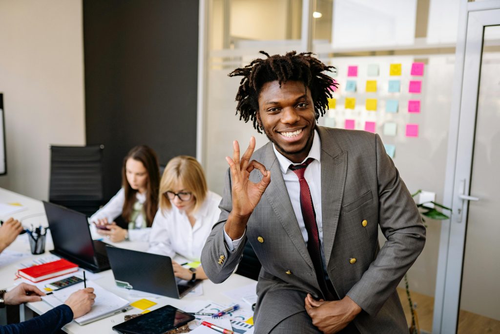 Im Büro: Zwei Mitarbeiterinnen arbeiten konzentriert im Hintergrund. Im Vordergrund sitzt ein fröhlicher Mitarbeiter, der seine Zufriedenheit mit einer positiven Handgeste zeigt.