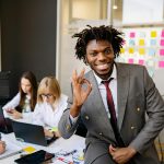 Im Büro: Zwei Mitarbeiterinnen arbeiten konzentriert im Hintergrund. Im Vordergrund sitzt ein fröhlicher Mitarbeiter, der seine Zufriedenheit mit einer positiven Handgeste zeigt.