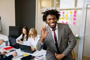 Im Büro: Zwei Mitarbeiterinnen arbeiten konzentriert im Hintergrund. Im Vordergrund sitzt ein fröhlicher Mitarbeiter, der seine Zufriedenheit mit einer positiven Handgeste zeigt.