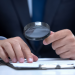 Person im Anzug prüft mit einer Lupe ein Dokument auf einem Schreibtisch – Symbol für Background Check und sorgfältige Überprüfung von Bewerbungsunterlagen.