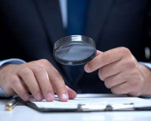 Person im Anzug prüft mit einer Lupe ein Dokument auf einem Schreibtisch – Symbol für Background Check und sorgfältige Überprüfung von Bewerbungsunterlagen.