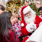 Der Weihnachtsmann in rotem Kostüm mit weißem Bart sitzt vor einem geschmückten Weihnachtsbaum und zeigt auf eine Liste. Um ihn herum stehen mehrere Kinder in festlicher Kleidung, die gespannt zuhören und lächeln.