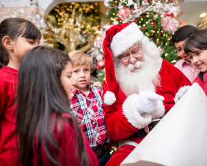 Der Weihnachtsmann in rotem Kostüm mit weißem Bart sitzt vor einem geschmückten Weihnachtsbaum und zeigt auf eine Liste. Um ihn herum stehen mehrere Kinder in festlicher Kleidung, die gespannt zuhören und lächeln.