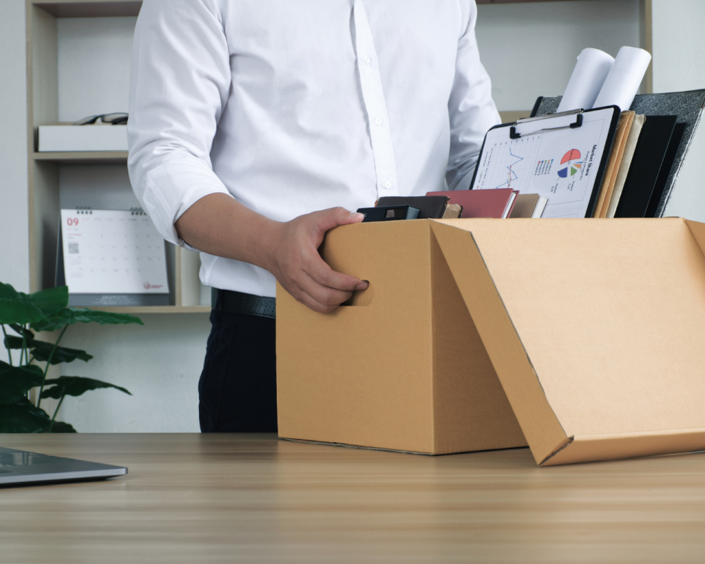 Eine Person im weißen Hemd räumt ihren Arbeitsplatz und hält eine braune Kartonschachtel mit Unterlagen, Ordnern und einem Klemmbrett, auf dem Diagramme zu sehen sind; im Hintergrund stehen ein Regal, ein Kalender und eine Zimmerpflanze.