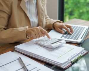 Person berechnet Kosten mit einem Taschenrechner am Schreibtisch, neben einem Laptop und Unterlagen mit Zahlen und Diagrammen.