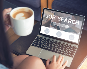 Person sitzt mit Kaffee auf dem Sofa und sucht auf einem Laptop nach Jobs („Job Search“ auf dem Bildschirm).